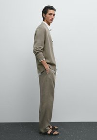 Beiger Cardigan mit strukturiertem Strick, über einem weißen Hemd getragen. Helle, locker sitzende Stoffhosen und dunkle Sandalen vervollständigen das Outfit.