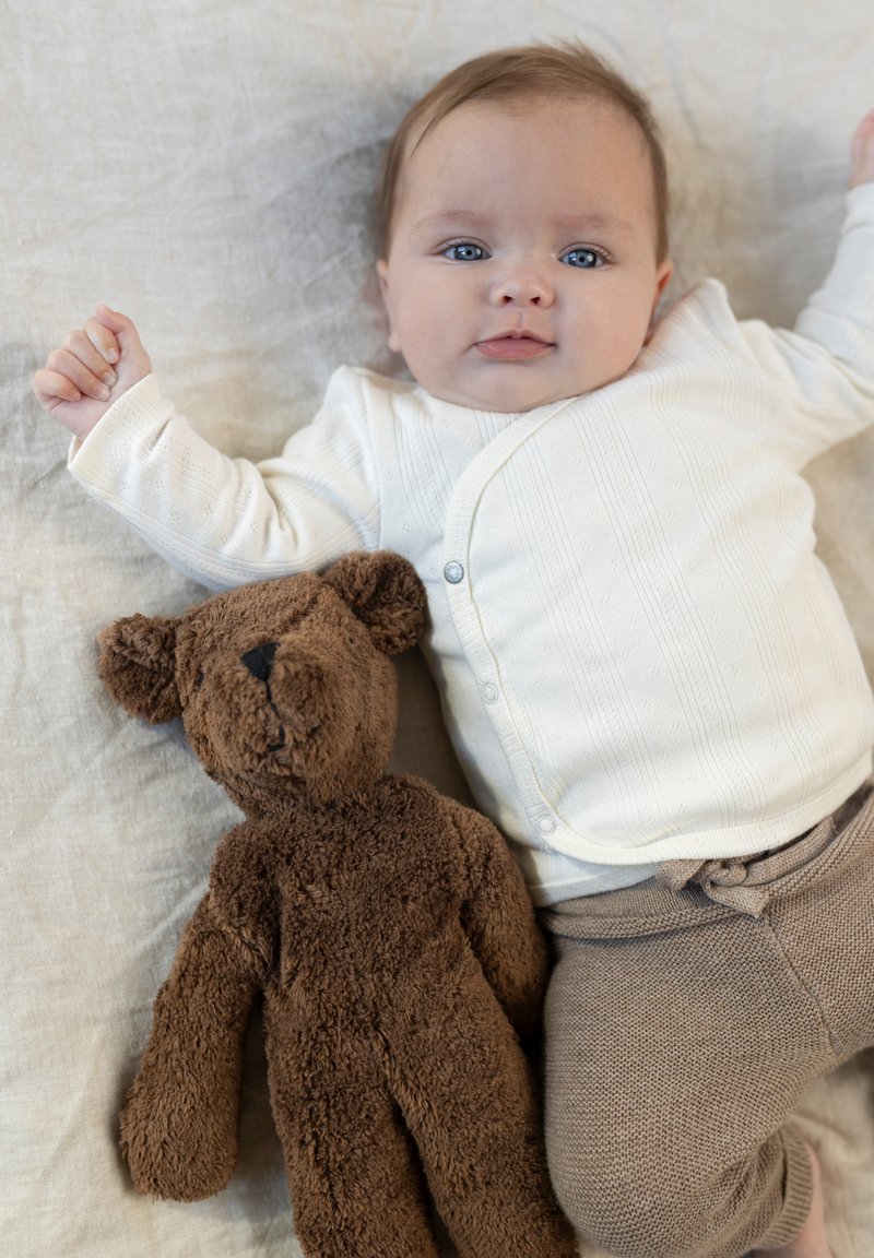 Baby mit blauen Augen, das auf einer hellen Oberfläche liegt, trägt eine weiße Strickjacke und beige Hose, neben einem braunen Plüschbären.