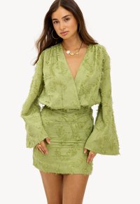 Robe mini verte avec des manches longues évasées et un tissu texturé, présentant un décolleté en V profond et des détails effilochés doux sur l'ensemble.