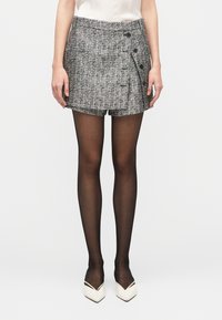 Pantalones cortos de tweed gris con diseño envolvente, que cuentan con bolsillos laterales y botones negros. Combinados con medias negras y zapatos blancos puntiagudos.