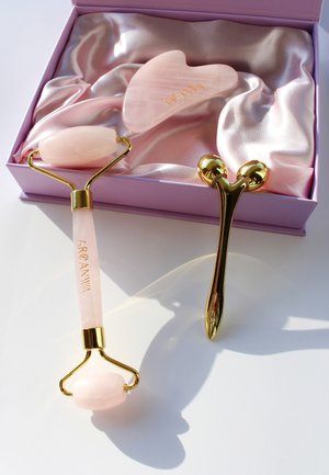 Rouleau facial en quartz rose et outil gua sha dans une boîte violette avec un revêtement en satin. Accents dorés, texture lisse, formes courbées et gravure de la marque.