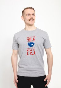Grå bomulds t-shirt med rød og blå tekst, et blåt ko-grafik, og sætningerne "LAVET MED MÆLK" og "SIDEN 1921." Korte ærmer, rund hals.