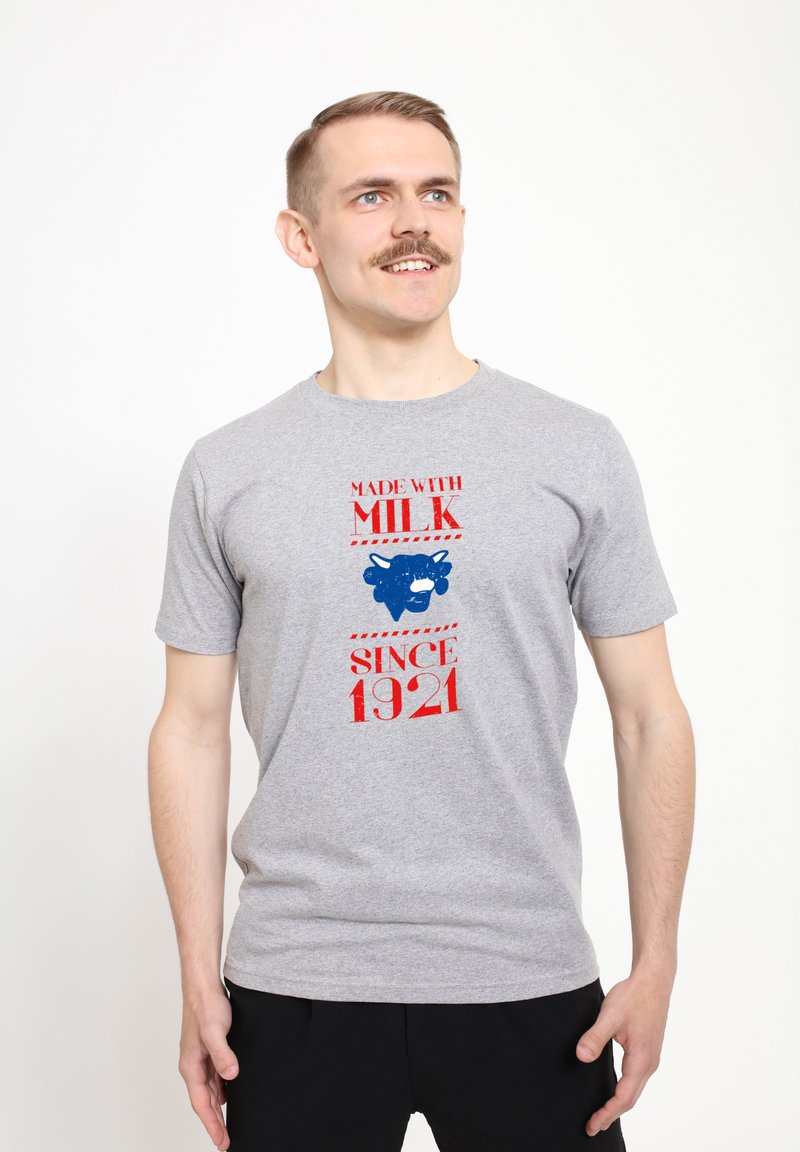 Grå bomulds t-shirt med rød og blå tekst, et blåt ko-grafik, og sætningerne "LAVET MED MÆLK" og "SIDEN 1921." Korte ærmer, rund hals.
