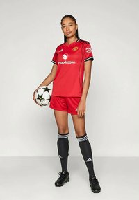 Equipamento de futebol vermelho do Manchester United, apresentando uma camisola de manga curta com detalhes em preto, combinada com calções vermelhos, meias pretas e botas.