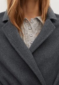 Col de manteau en laine grise sur un pull beige à boutons en maille torsadée porté par une personne aux longs cheveux châtain clair.