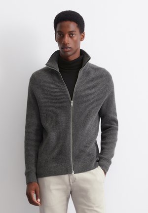 Grauer, gerippter Reißverschluss-Pullover mit hohem Kragen, der eine glatte Textur und silberne Reißverschlussdetails aufweist, kombiniert mit einem schwarzen Rollkragenpullover darunter.
