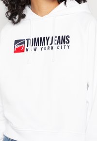 Sudadera blanca con cordones que presenta un logo de Tommy Jeans y el texto "New York City" en la parte delantera, llevada por una persona con cabello rizado.