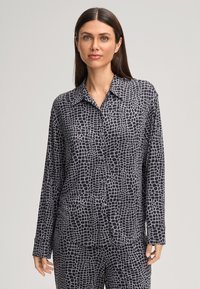 Een vrouw in een zwart en grijs geblokt overhemd met lange mouwen en bijpassende broek, staand tegen een effen lichte achtergrond.