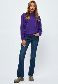 Lila tröja med puffärmar, i kombination med blå utsvängda jeans. Skorna är beige loafers med tjock sula. Bakgrunden är enkel vit.