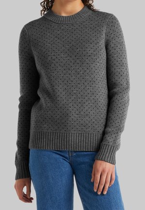 Woman wearing a fitted grey knit sweater with small black dots and ribbed cuffs and hem, paired with blue jeans.
