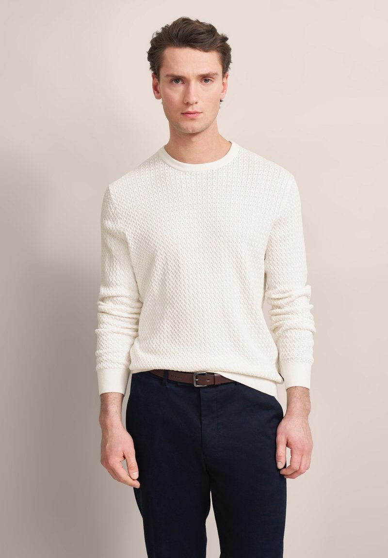 Cremefarbener gestrickter Pullover mit einem strukturierten Muster und rundem Ausschnitt, kombiniert mit dunklen Hosen und einem braunen Gürtel.