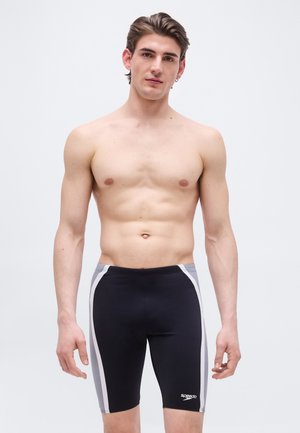 Junger Mann steht vor hellem Hintergrund, trägt schwarze und graue Speedo-Schwimmjammers, ist oben ohne und zeigt einen neutralen Gesichtsausdruck.