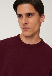 Maglione in jersey bordeaux con collo rotondo e discreto dettaglio del logo. Tessuto morbido e design aderente, che mette in risalto una silhouette pulita.