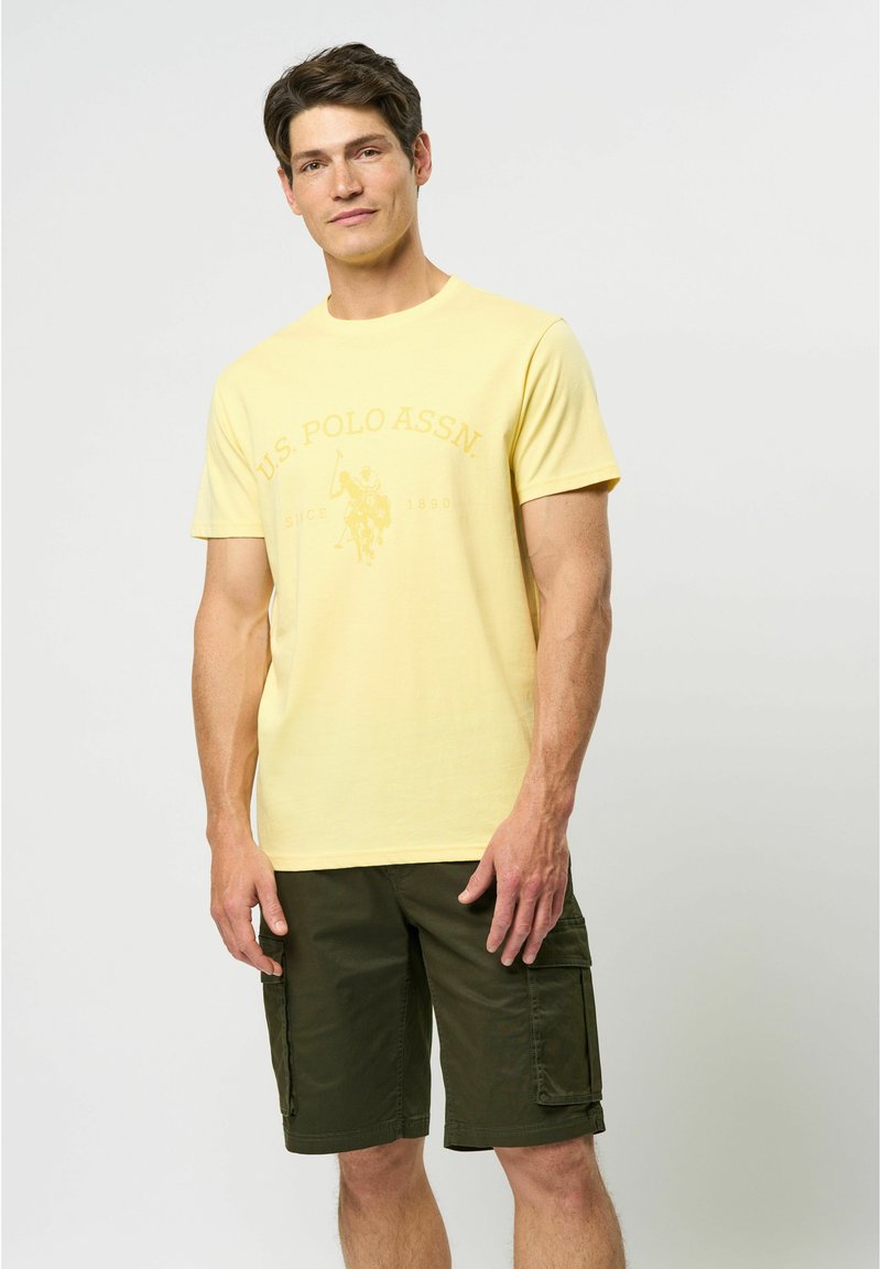 Tricou din bumbac galben cu logo-ul U.S. Polo Assn. imprimat. Asortat cu pantaloni scurți cargo de culoare verde închis. Design simplu cu guler tip crew.
