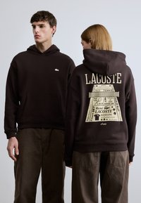 Καφέ φούτερ με κουκούλα και μπροστινή τσέπη, με μικρό λογότυπο Lacoste. Στο πίσω μέρος υπάρχει γραφική εκτύπωση με κείμενο και σχέδια που σχετίζονται με το τένις.