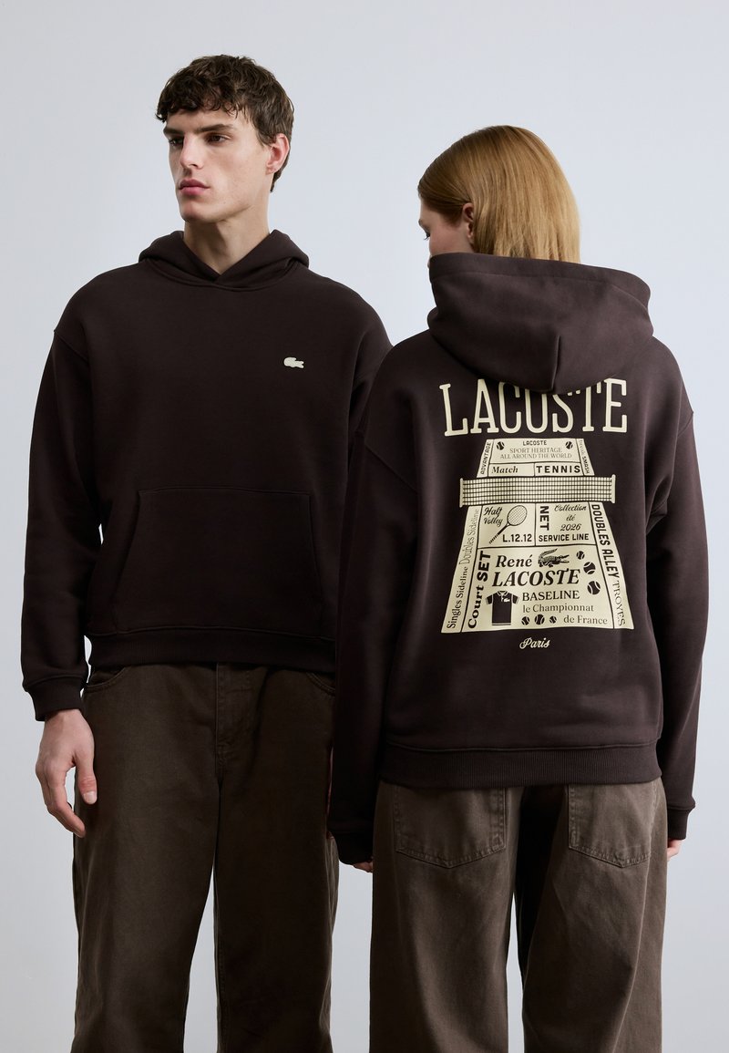 Καφέ φούτερ με κουκούλα και μπροστινή τσέπη, με μικρό λογότυπο Lacoste. Στο πίσω μέρος υπάρχει γραφική εκτύπωση με κείμενο και σχέδια που σχετίζονται με το τένις.