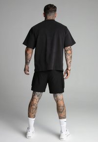 Camiseta negra de manga corta con un corte relajado, combinada con pantalones cortos cargo negros. El modelo lleva zapatillas blancas y tatuajes en las piernas con patrones visibles.