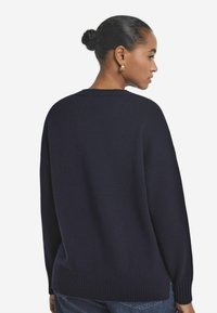 Marineblaue Strickjacke mit Rundhalsausschnitt, fallenden Schultern und Rippensaum. Weicher Stoff und lässige Passform, kombiniert mit Jeans.