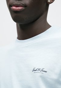 Ανοιχτό μπλε βαμβακερό φούτερ με στρογγυλή λαιμόκοψη και κοντά μανίκια. Στο αριστερό στήθος εμφανίζεται η φράση «Jack & Jones Archive Selection» σε σκούρο μπλε χρώμα.