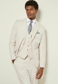 Costume trois pièces beige clair avec une veste ajustée, un gilet et un pantalon sur mesure. Une cravate texturée et un mouchoir de poche à motifs ajoutent du détail.