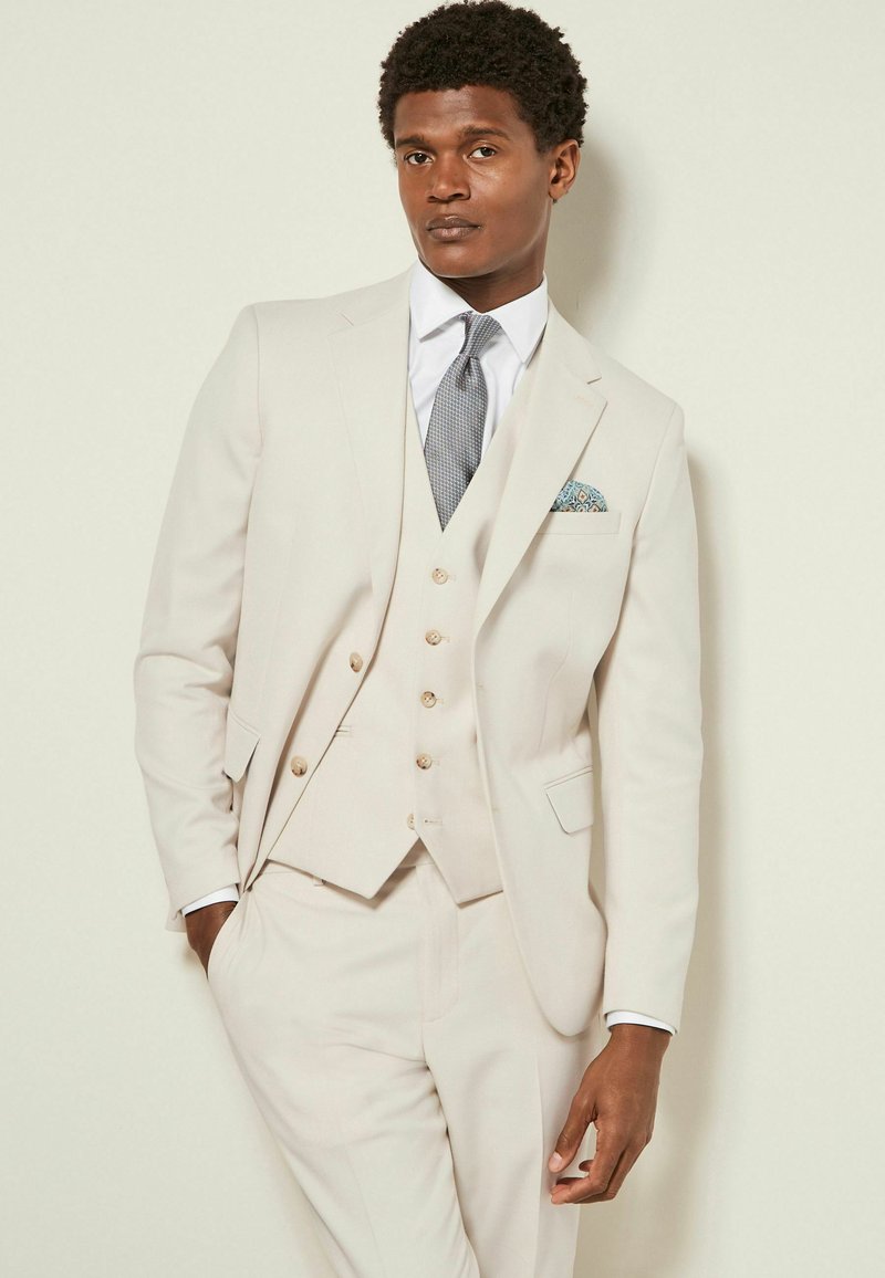 Costume trois pièces beige clair avec une veste ajustée, un gilet et un pantalon sur mesure. Une cravate texturée et un mouchoir de poche à motifs ajoutent du détail.