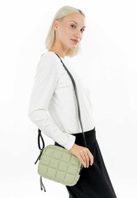 Bolso bandolera acolchado verde hecho de material texturizado, con cierre de cremallera y una larga correa negra; combinado con una blusa de manga larga blanca.