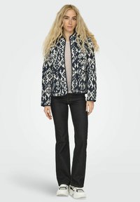 Giacca zip-up a righe blu navy e bianca con colletto alto, abbinata a un maglione chiaro, jeans scuri e scarpe da ginnastica bianche.