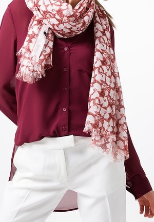 Femme portant un chemisier bordeaux à boutons, un pantalon blanc et une écharpe blanche avec des motifs floraux rouges et des bords frangés.