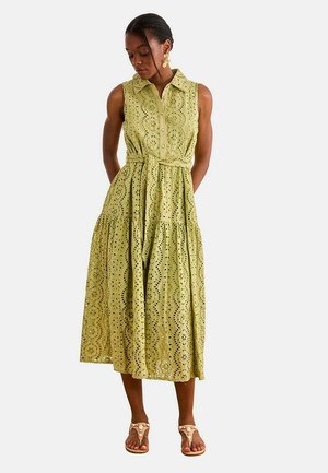 Mujer vestida con vestido midi sin mangas en verde claro con ojales, con cuello y lazo en la cintura, combinado con sandalias beige y pendientes dorados con diseño de flores.