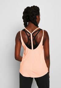 Top sin mangas color durazno con un diseño cruzado de tirantes negros en la parte posterior. Textura suave y dobladillo redondeado, superpuesto a un sujetador deportivo negro.