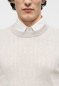 Άνδρας που φοράει ανοιχτό μπεζ πουλόβερ με πλεκτό μοτίβο πάνω από λευκό πουκάμισο με γιακά, εμφανίζοντας το κάτω μέρος του προσώπου και το πάνω μέρος του στήθους.