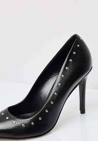 Pantof cu toc înalt din piele neagră, cu vârf ascuțit și nasturi decorativi argintii pe margine, având o textură netedă și un design elegant.