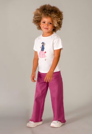 Bambino con capelli ricci che indossa una maglietta bianca con grafica, pantaloni viola larghi e scarpe da ginnastica bianche, in piedi davanti a uno sfondo semplice.