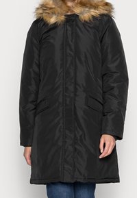 Parka noire matelassée avec une capuche bordée de fourrure synthétique, dotée de poches latérales et d'une silhouette droite, fabriquée à partir d'un tissu lisse et brillant.