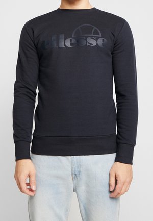 Person wearing a black long-sleeve sweatshirt with dark logo text on the chest and light blue jeans, standing against a plain background.