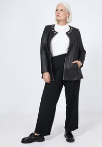 Veste en simili cuir noire avec des clous argentés et des accessoires ronds, associée à une chemise blanche et un pantalon large noir. Des chaussures noires complètent le look.