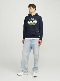 Sudadera navy con logotipo frontal, capucha con cordones y una capa blanca debajo. Combinada con jeans de corte holgado en azul claro y zapatillas blancas.