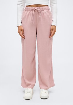 Kobieta ubrana w jasnoróżowe spodnie o szerokich nogawkach z elastyczną talią i sznurkiem, do tego białe trampki i krótka biała bluzka z długim rękawem.