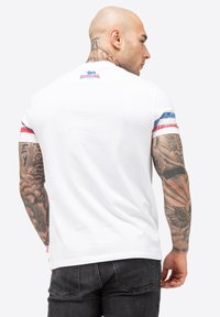 Balta medvilninė T-shirt marškiniai su mėlynomis ir raudonomis dryžuotomis akcentais ant rankovių. Ant nugaros yra logotipas, o ant rankos – sudėtingos tatuiruočių detalės.