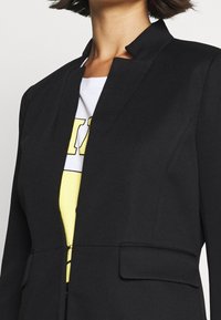 Svart blazer med en strukturerad design, med revere krage, två fickor och textil med struktur. Bärs över en vit och gul skjorta.