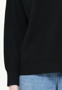 Pull noir en maille avec des manches longues, doté d'une bordure et de poignets côtelés, associé à un jean en denim bleu clair. La texture semble douce.