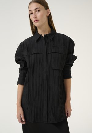 Fekete, textúrált ing, gombos elülső résszel, két mellzsebbel és felgöngyölt ujjal. Kissé túlságosan oversized fazonnal és gallérral rendelkezik.