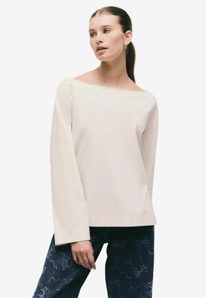 Helles beige Langarmtop mit weitem Bootsausschnitt, aus glattem Stoff, kombiniert mit dunklen Jeans, die ein dezentes Schleifenmuster aufweisen.
