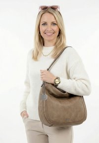 Blonde woman in white sweater and beige pants smiling, holding a taupe shoulder bag with braided trim and wearing a gold watch and pink sunglasses.