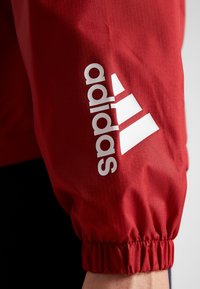 Červená sportovní bunda s texturovaným povrchem, elastickými manžetami a výrazným bílým logem Adidas se třemi pruhy.