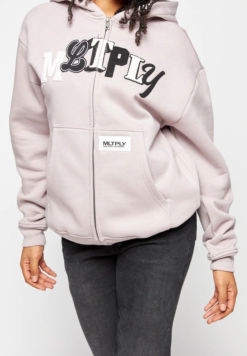 Reißverschluss-Hoodie in Hellrosa mit schwarz-weißer Aufschrift "MLTPLY". Besitzt Fronttaschen, gerippte Bündchen und einen strukturierten Stoff.