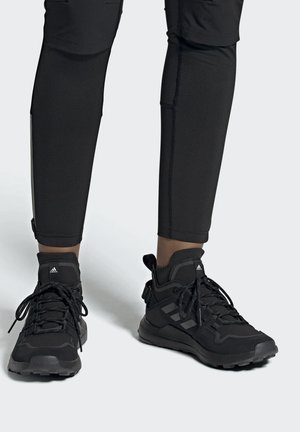 Czarne wysokie buty sportowe Adidas z sznurowadłami i logo, noszone z czarnymi legginsami, pokazane z przodu na białej podłodze.