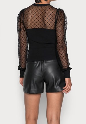 Femme portant un haut en dentelle noire transparente à manches longues avec des épaules bouffantes et un short en cuir noir, vue de dos sur fond uni.