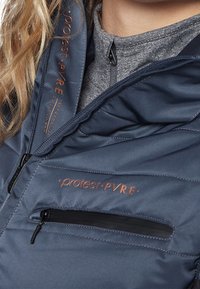 Nahaufnahme einer Person, die eine navyblaue Jacke mit orangefarbenen "protest PVRE" Logos und einer grauen zip-up Innenlage trägt.