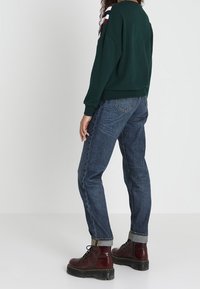 Sudadera verde con hombros caídos, rayas blancas y burdeos en las mangas, combinada con unos vaqueros de denim azul oscuro y botas burdeos con cordones.